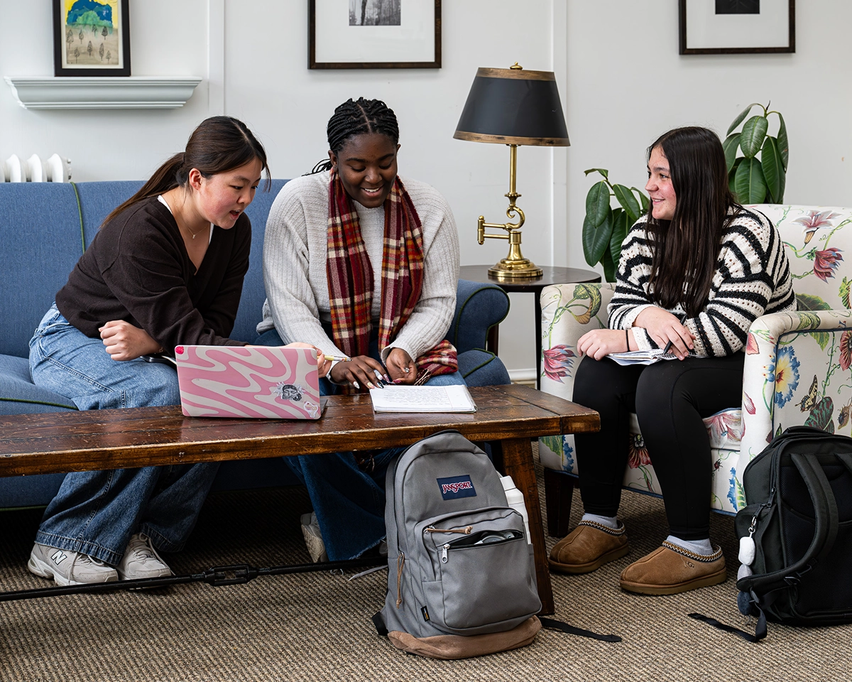 Three young women sit together, smiling and working at a coffee table with papers and a laptop in a cozy room with couches, houseplants, and a lamp. Backpacks rest on the floor nearby.