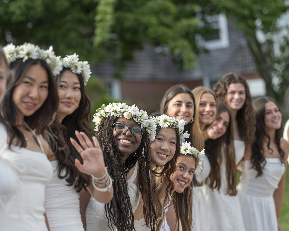 A group of young women in white dresses and flower crowns smile and pose outdoors, with one waving at the camera. Trees and a building are visible in the background.