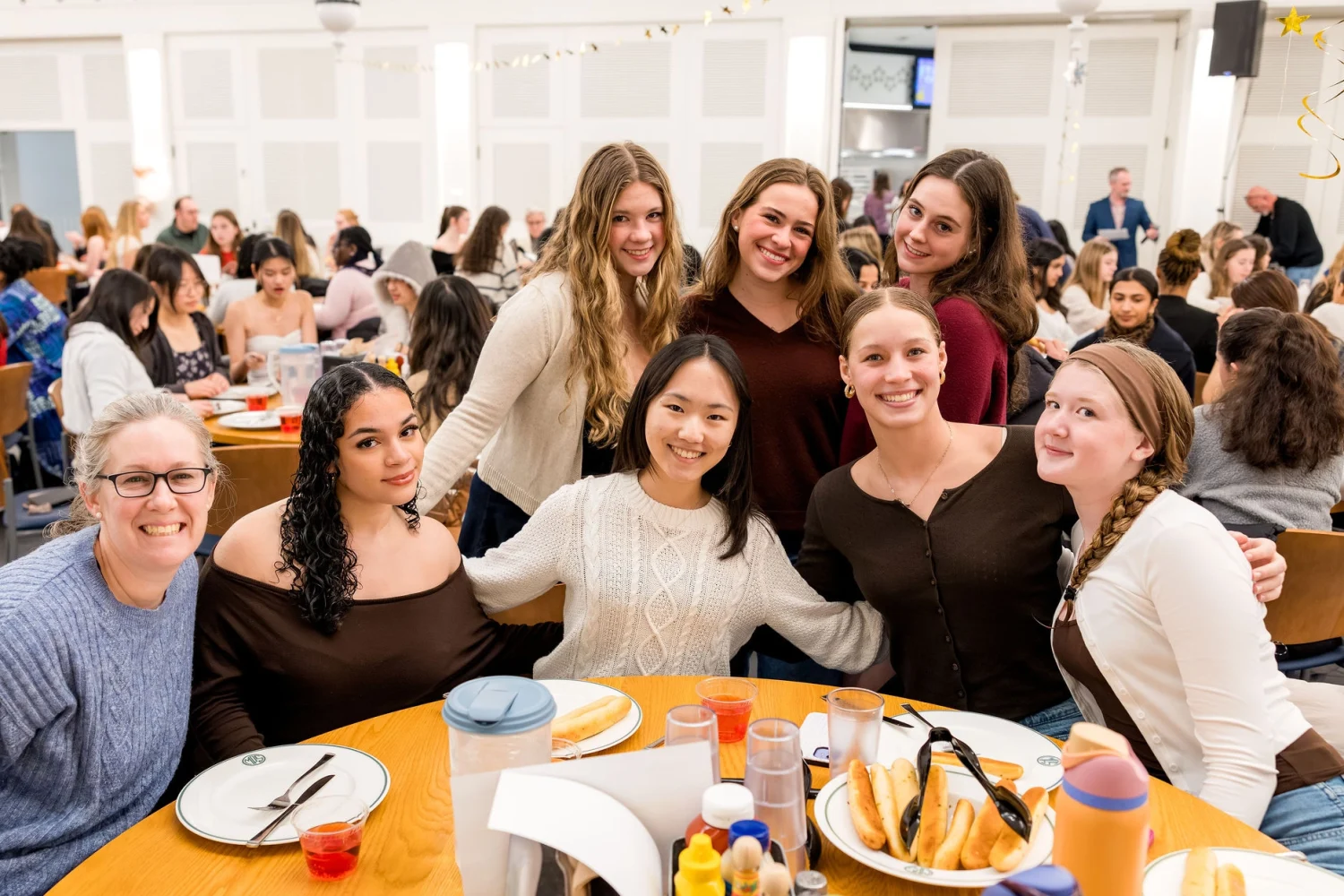 A group of seven women smile and pose together around a table with food in a bright, busy dining hall filled with other people.
