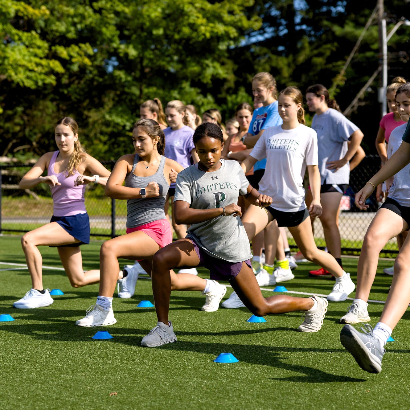 A group of teens exercise outdoors on a field, performing lunges in front of blue cones under the sun, with trees and a fence in the background.