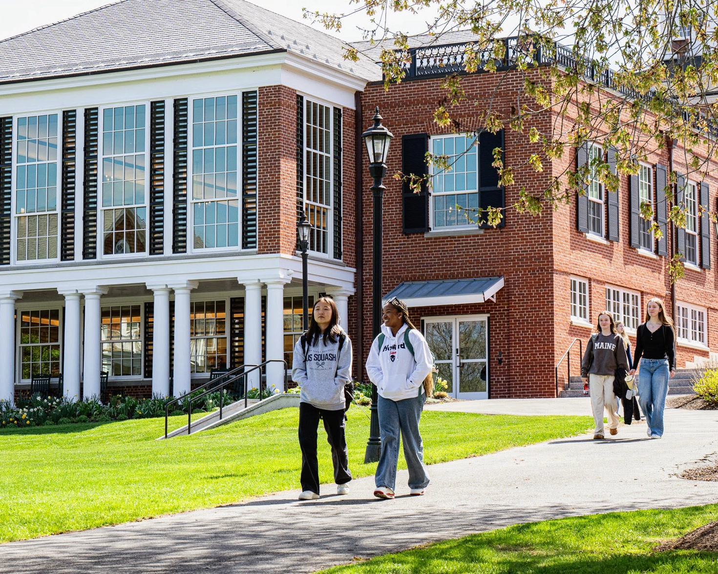 Students walk on a sunny path in front of a large brick and white academic building with tall windows. Some trees with green leaves are visible, and the sky is clear.