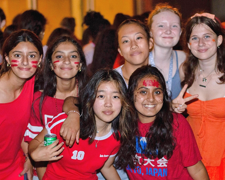A group of smiling teens wearing colorful clothes and face paint pose together at a lively event, some with peace signs and arms around each other, enjoying the festive atmosphere.