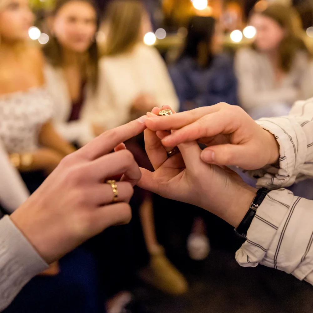 A person places a ring on anothers finger in a close-up, with a blurred group of people in the background suggesting a social gathering or celebration.