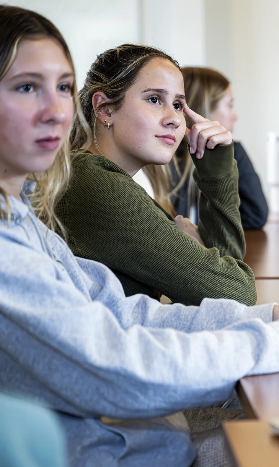 Three young people sit at a desk, listening attentively. The person in front wears a gray hoodie, the person behind wears a green sweater and rests their head on their hand, and the third person is blurred in the background.