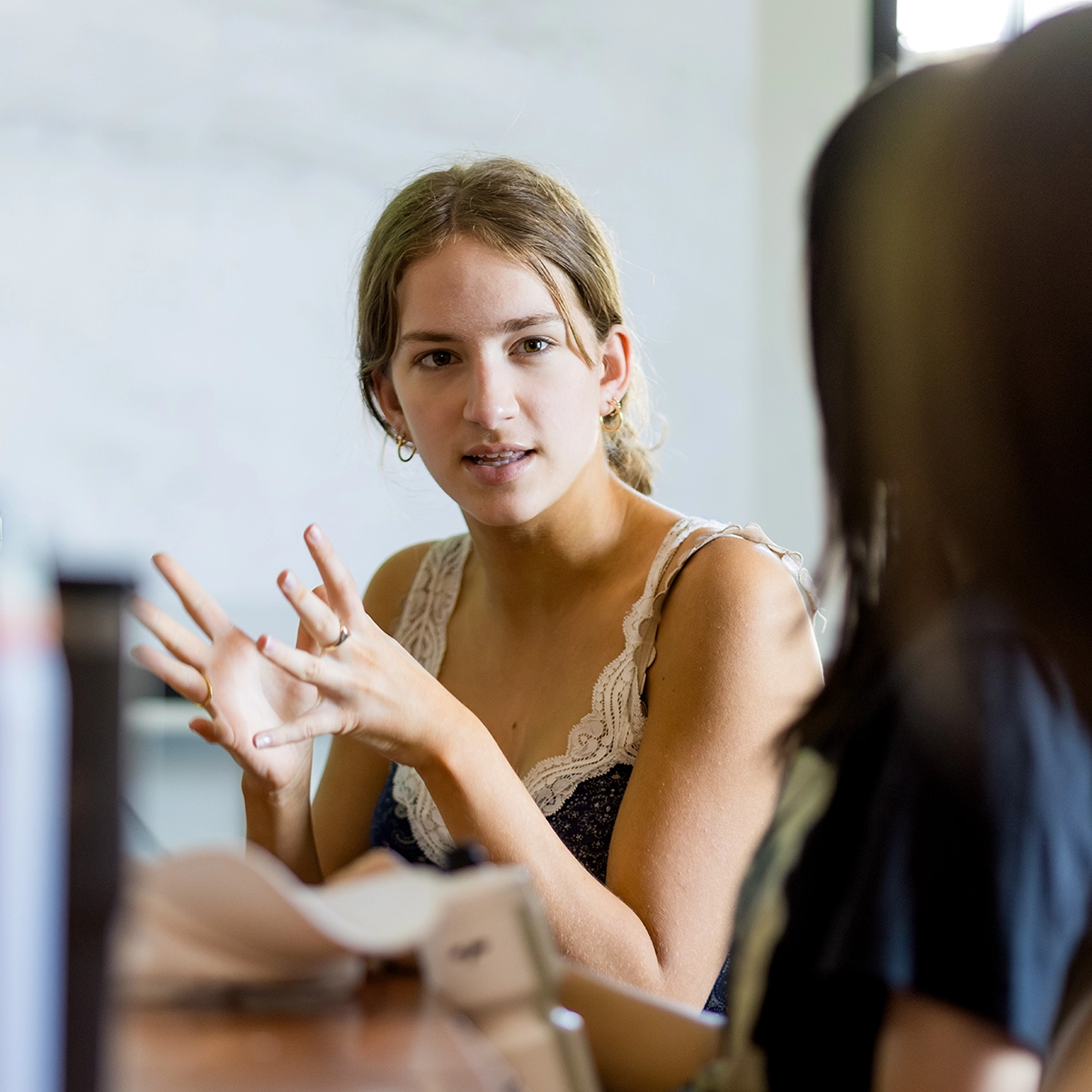 A young woman in a tank top gestures while speaking to another person, who is partially visible. They appear to be having a serious conversation indoors, with books or papers on the table.