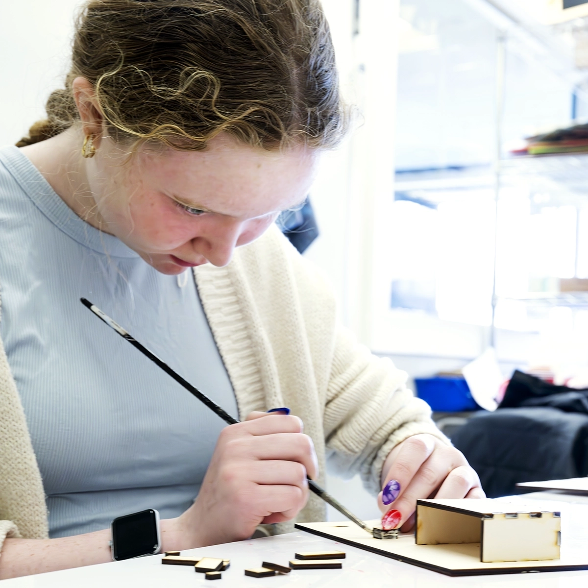 A person with curly hair and a cream sweater carefully paints small wooden pieces at a desk, focusing intently on their work in a bright room.