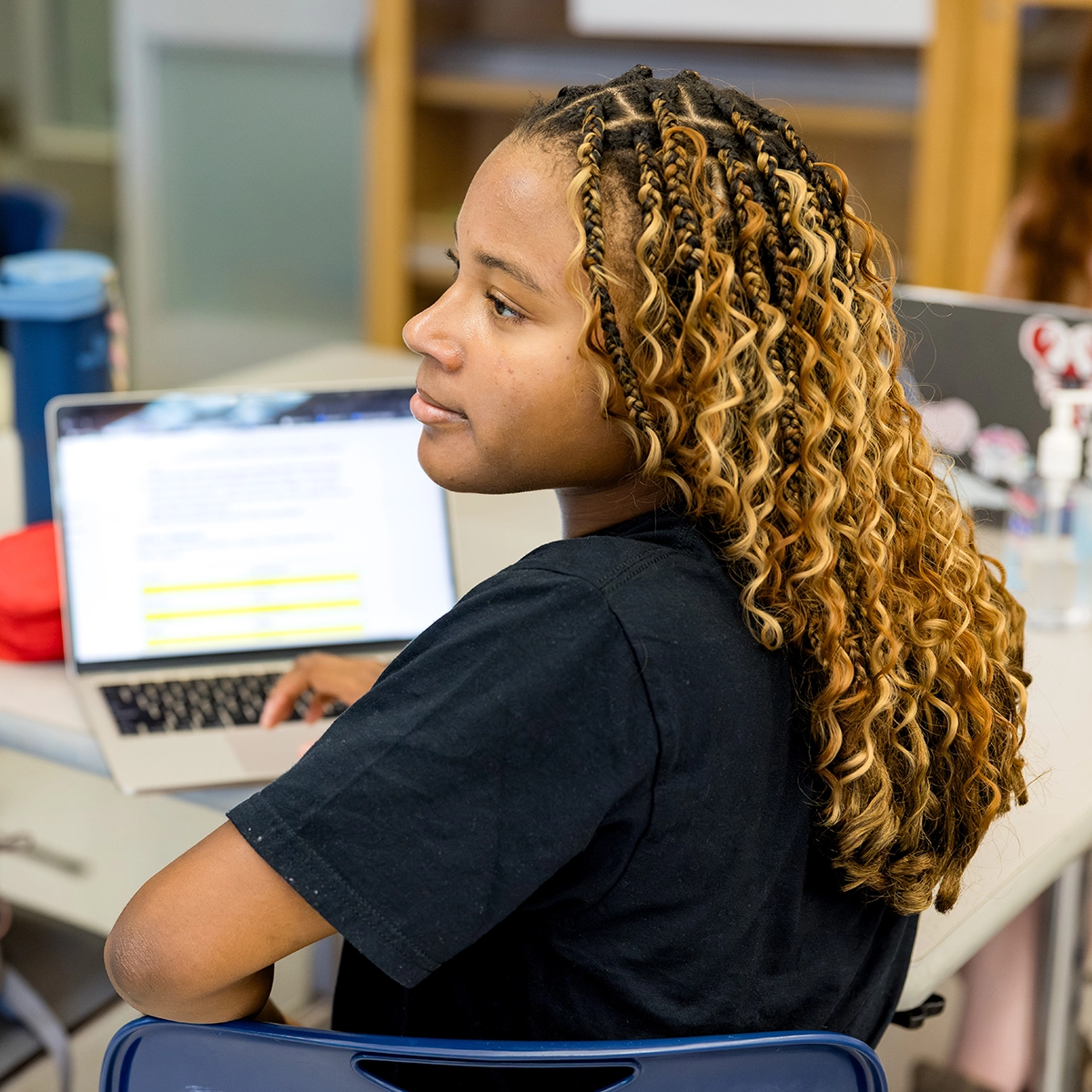 A student with curly, highlighted hair sits in a classroom, looking to the side. She is wearing a black shirt and is using a laptop with a highlighted document on the screen.