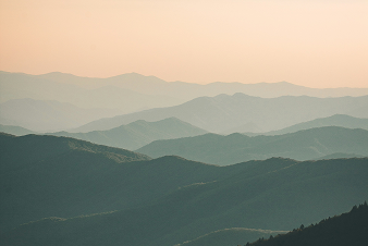 Softly lit mountain ridges stretch into the distance under a hazy, pastel sky, creating a layered effect with gentle gradients of green and blue.