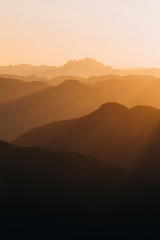 Golden sunlight shines over layers of mountain ridges at sunrise or sunset, creating a warm, hazy atmosphere with a prominent peak visible in the background.