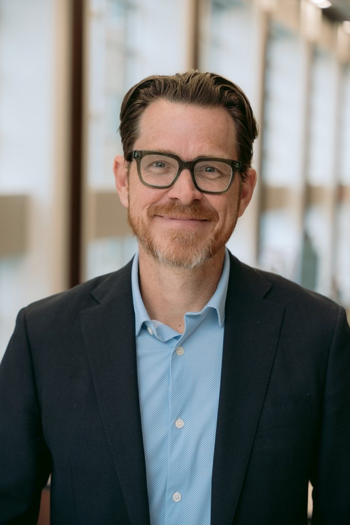 A man with brown hair, glasses, and a beard is smiling. He is wearing a blue dress shirt and a dark blazer, standing indoors with soft natural light and blurred windows in the background.