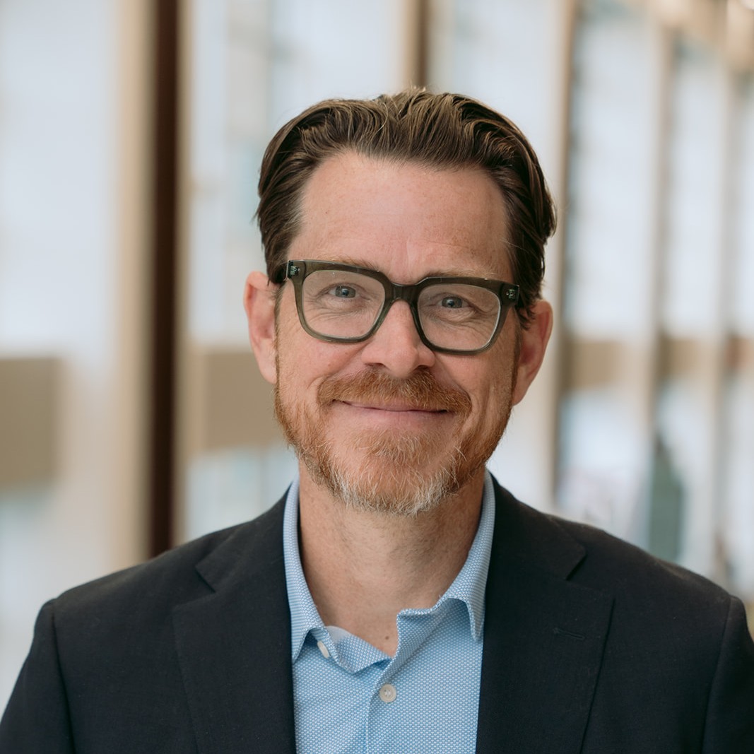 Man with short brown hair, beard, and glasses, wearing a dark blazer and light blue shirt, smiling in a brightly lit indoor setting with large windows in the background.