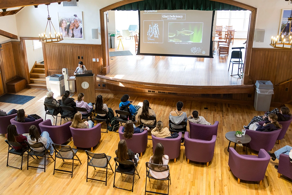 A person presents a science lecture on a stage to an audience seated in purple chairs in a spacious hall with wooden floors and large windows.