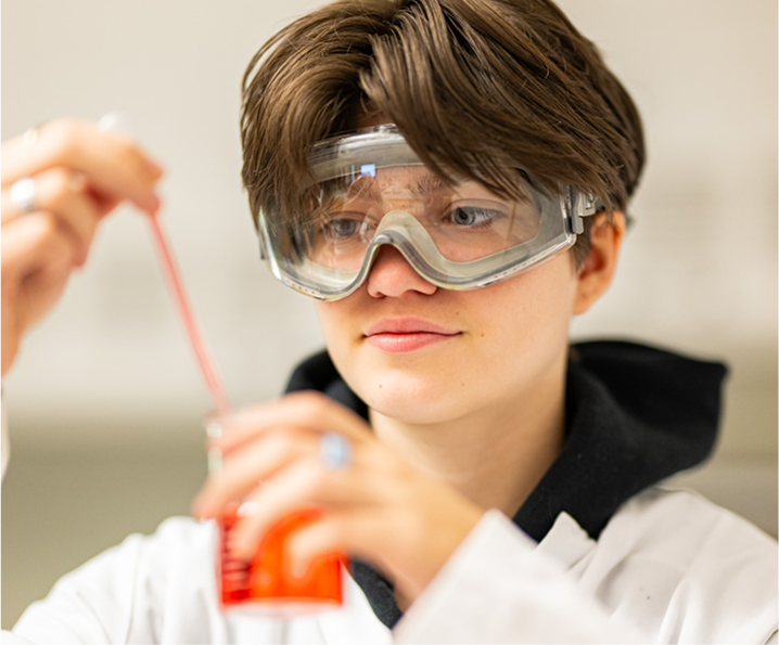 A person wearing safety goggles and a white lab coat uses a pipette to transfer liquid into a beaker filled with red solution in a laboratory setting.