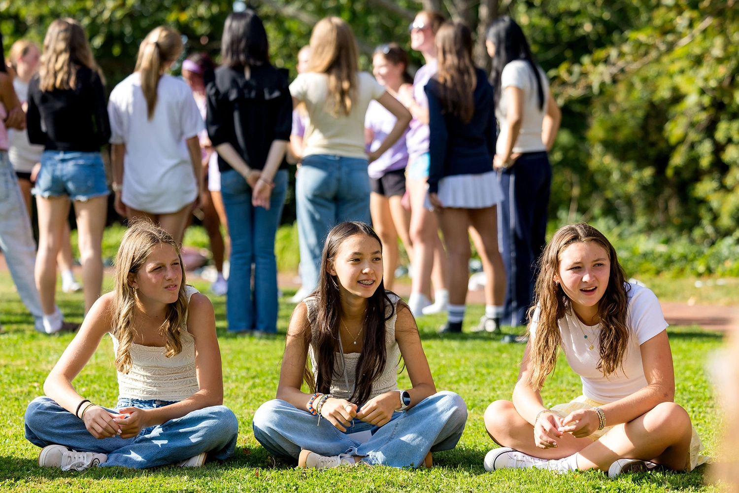 Three teenage girls sit on grass, smiling and talking, while a group of people stand and chat in the background under trees on a sunny day.