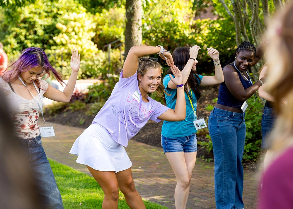 A group of young people stand outdoors on grass, smiling and dancing with arms raised. They wear casual summer clothes and name tags. Trees and sunlight are visible in the background.