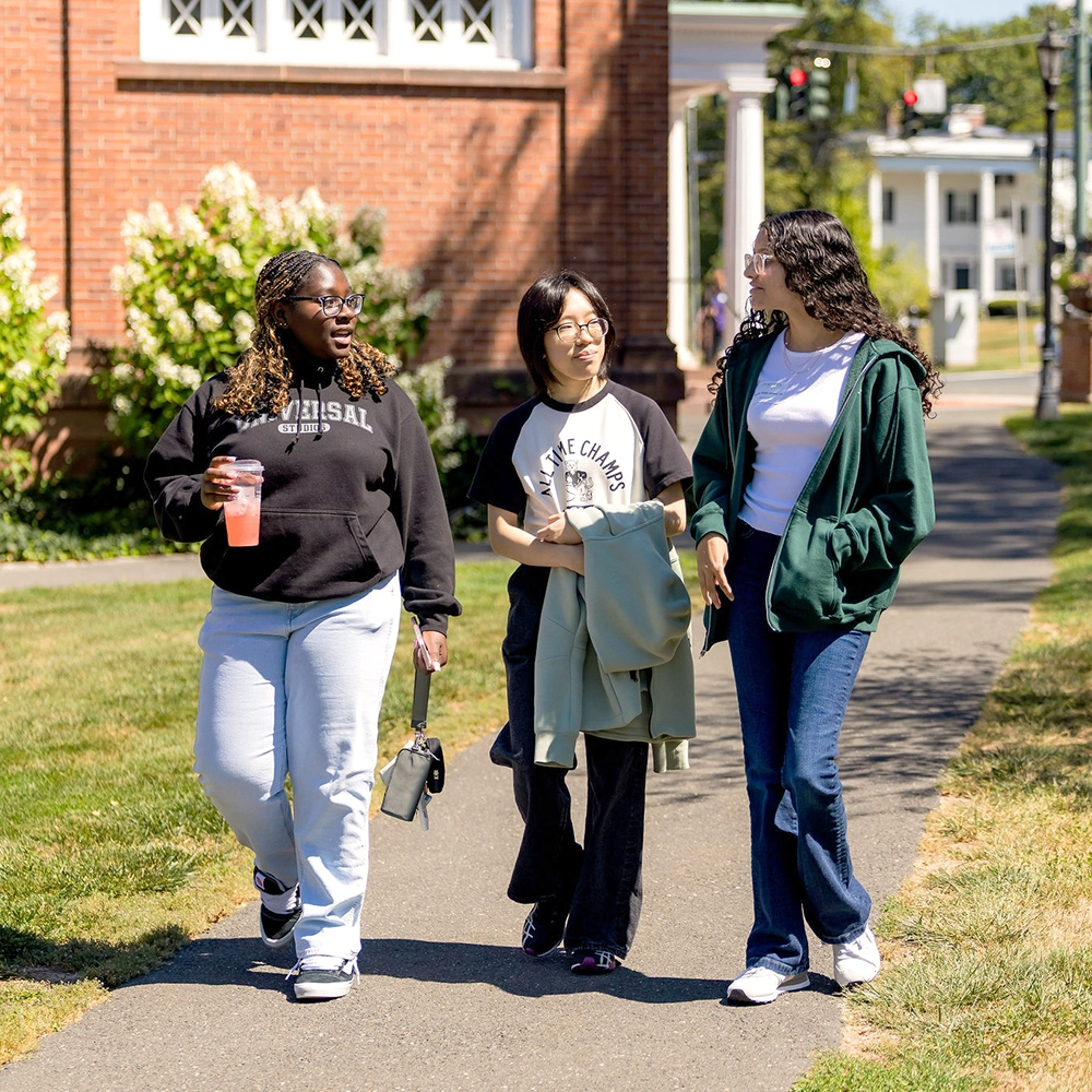 Three young women walk and talk on a campus sidewalk on a sunny day. One holds a drink, another carries a jacket, and all appear to be enjoying their time together.