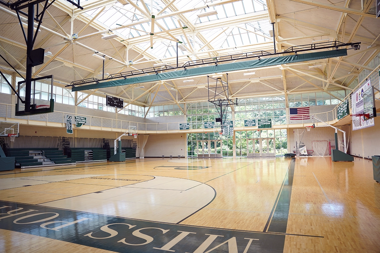 Empty indoor basketball court with wooden flooring, bleachers on the left, basketball hoops, banners, scoreboard, and large windows letting in natural light. An American flag is displayed on the far wall.