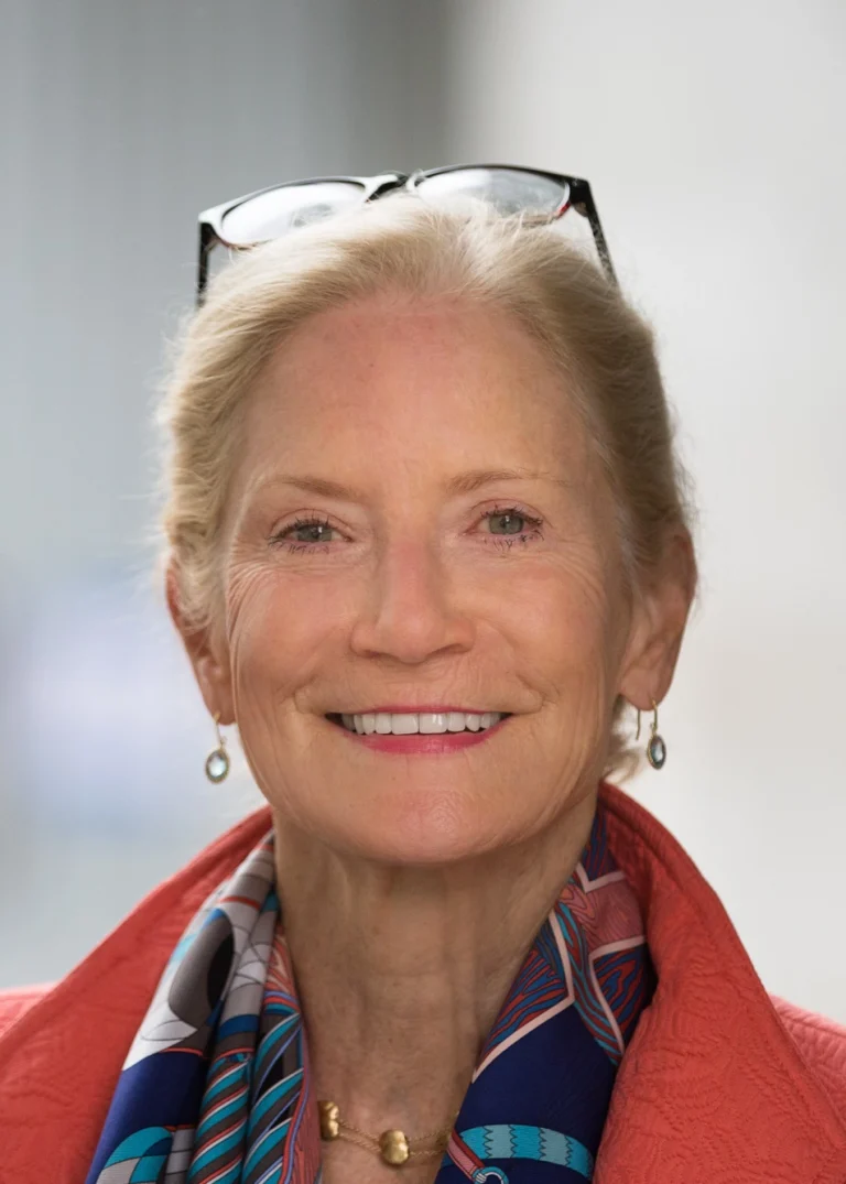 Smiling older woman with light skin, blond hair tied back, wearing glasses on her head, gold earrings, a colorful scarf, and a coral jacket, standing in front of a blurred, light background.