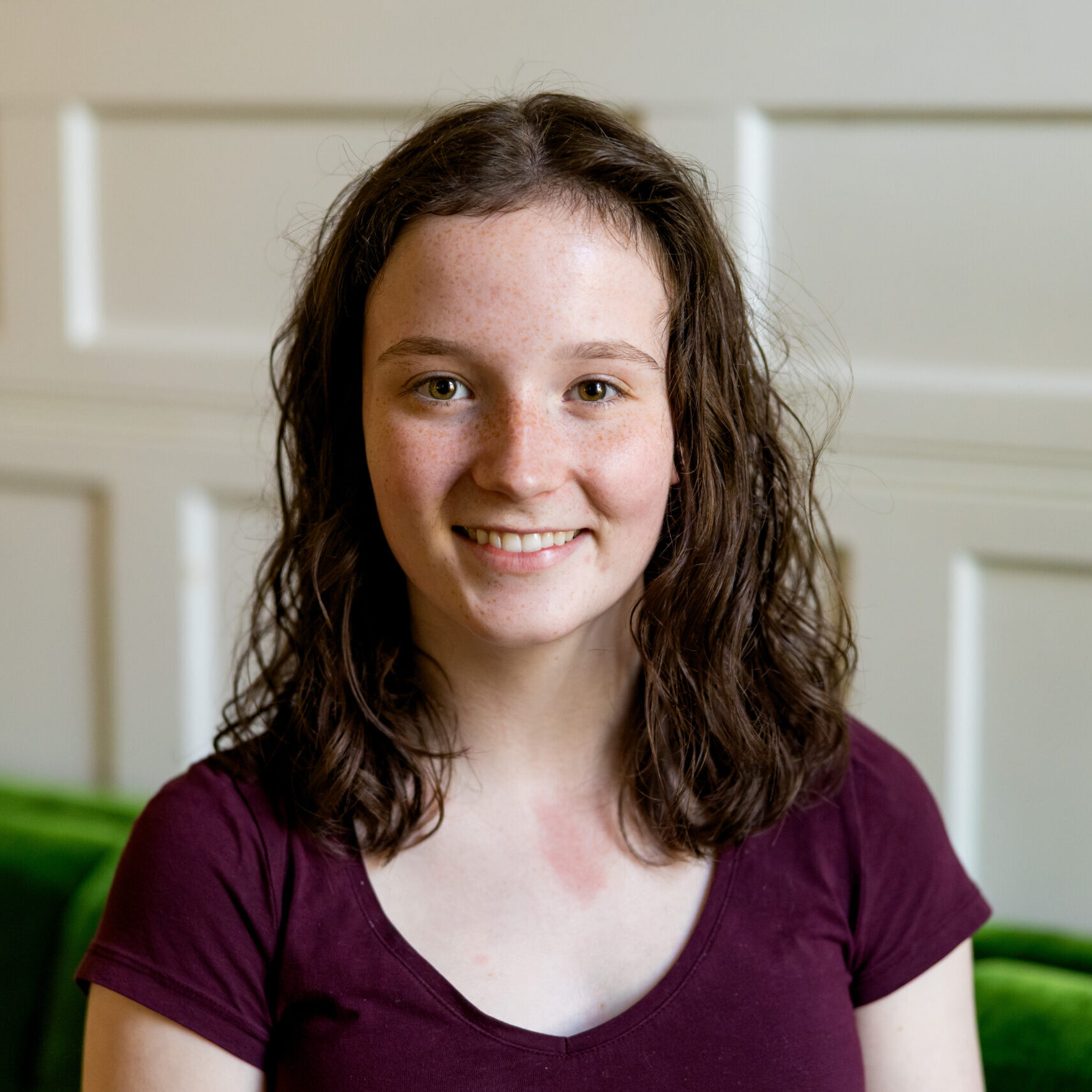 A young person with wavy brown hair and a maroon shirt smiles while sitting on a green couch in front of a white paneled wall.