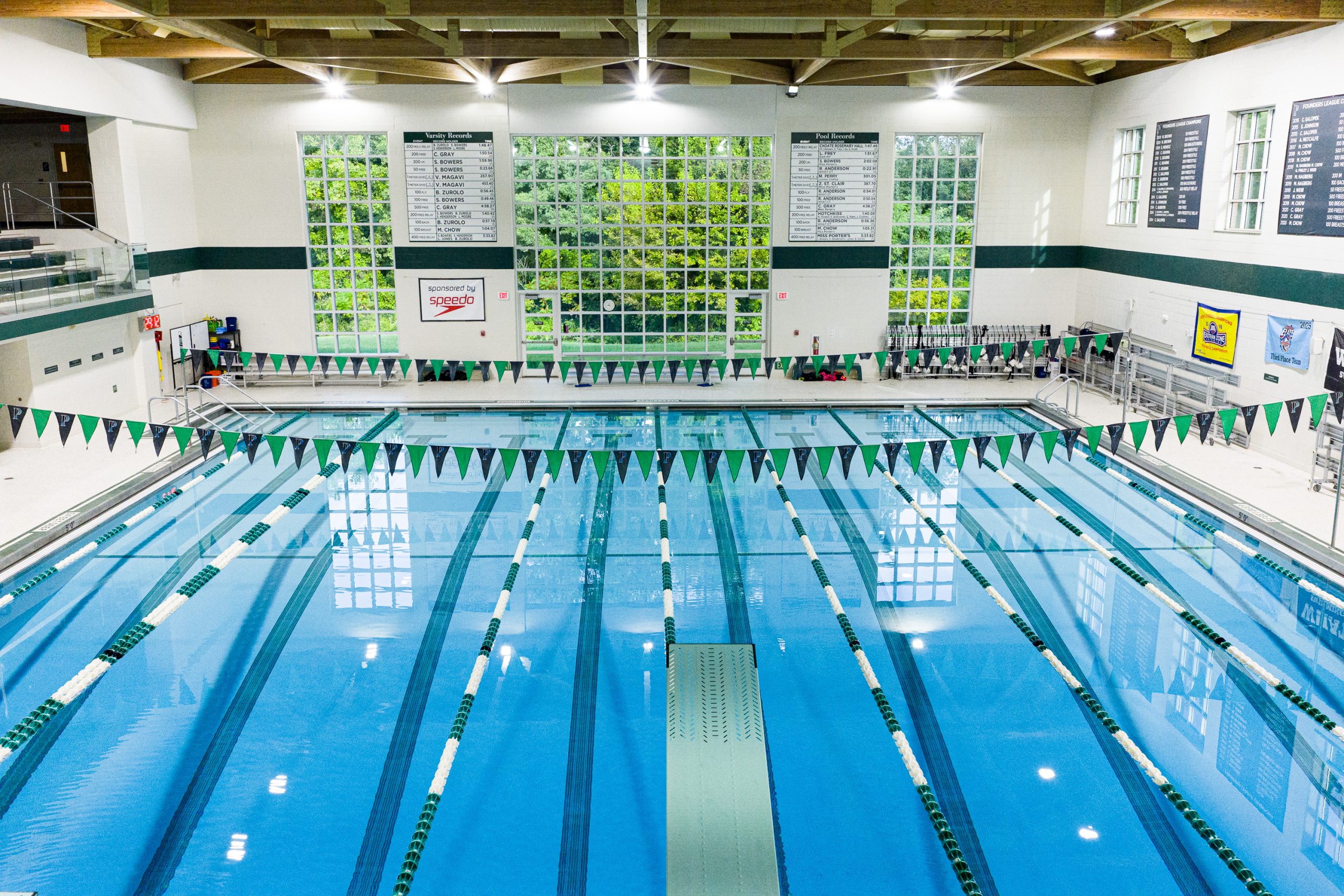 Porter's indoor swimming pool with multiple lanes, green and white lane dividers, and triangular flags above the water. Large windows and record boards line the white walls, and greenery is visible through the windows.
