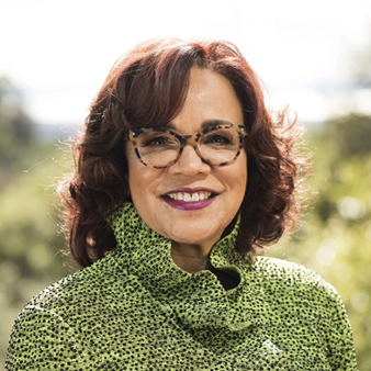 A woman with shoulder-length brown hair and glasses smiles outdoors, wearing a green patterned jacket. The background is blurred with natural light and greenery.