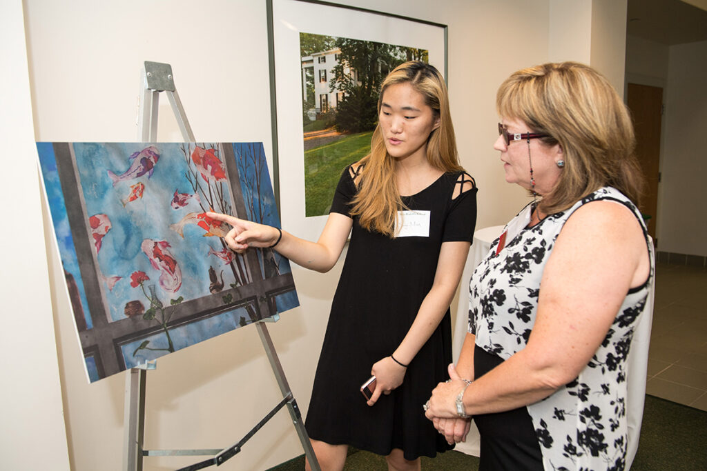 Two women stand next to an easel displaying a colorful painting of koi fish; one woman points at the artwork while explaining something to the other.