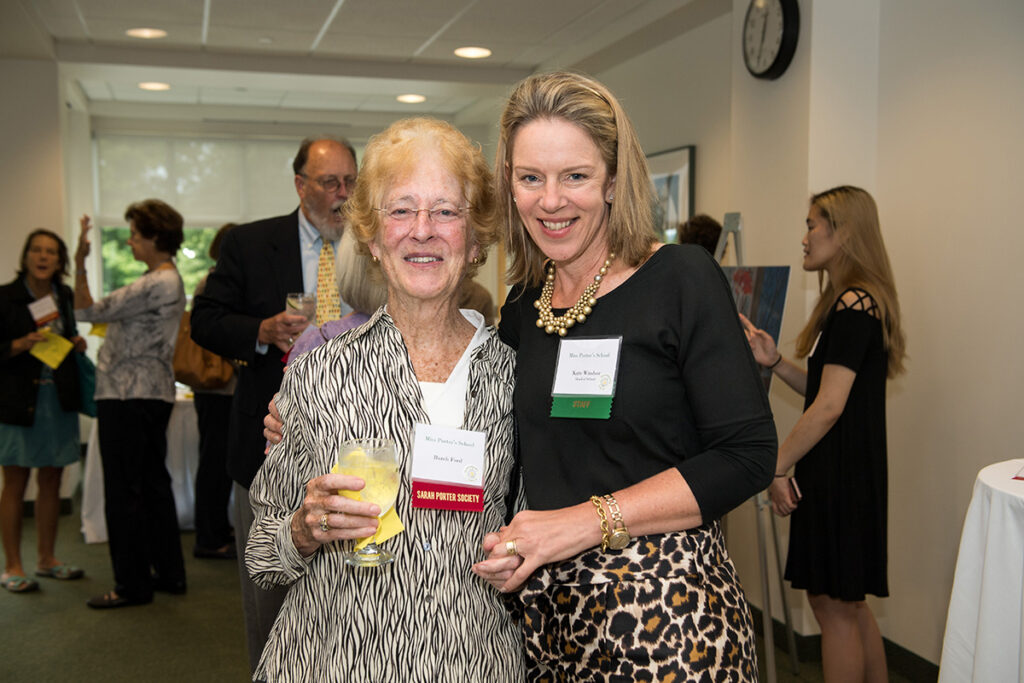Two women smiling and posing together at an indoor event; one holds a drink. Both wear name tags, and other people socializing can be seen in the background.
