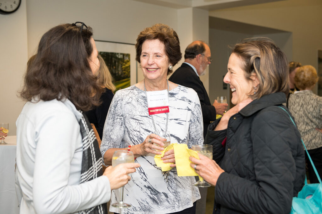 Three women stand together at a social event, smiling and holding drinks while talking. Other people mingle in the background.