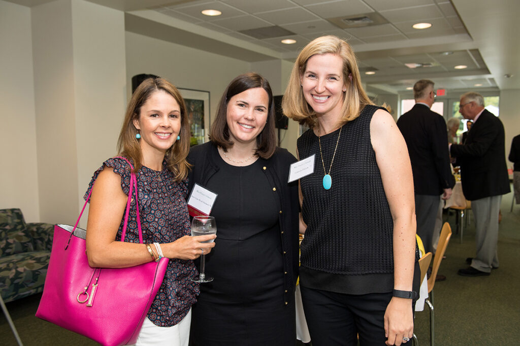 Three women smiling and posing together indoors at a social event. One woman holds a glass of water; another has a bright pink handbag. People talk in the background. The setting appears to be a reception or gathering.