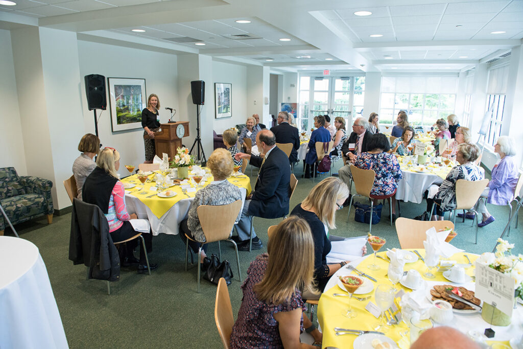 A woman speaks at a podium to a group of people seated at round tables covered with yellow tablecloths during a daytime event in a bright room.