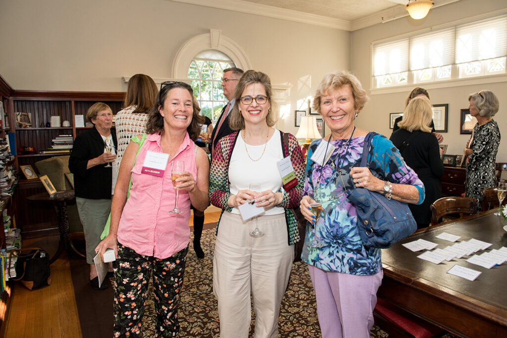 Three women smile at the camera at a social gathering in a well-lit room. They hold drinks and wear name tags. Other people are mingling in the background near bookshelves and tables.