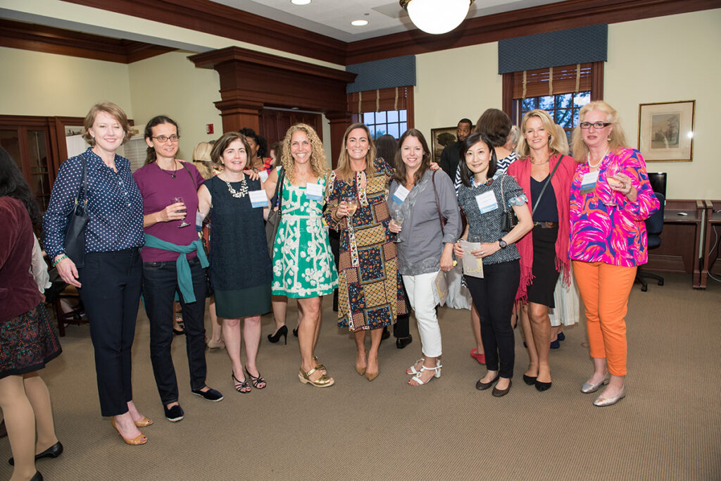Nine women stand together in a well-lit room, smiling at the camera. Most are holding drinks and wearing a variety of business casual and colorful outfits. Several people are mingling in the background.
