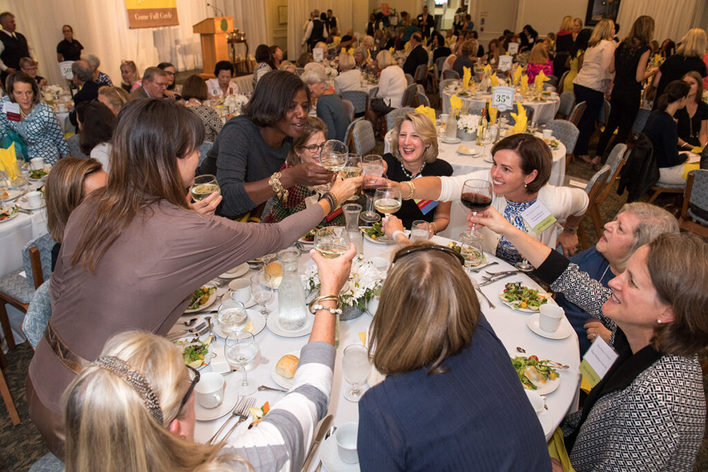 A group of women sitting around a banquet table raise their glasses in a toast at a crowded, elegant event. Other guests are visible in the background, with plates of salad and bread on the tables.