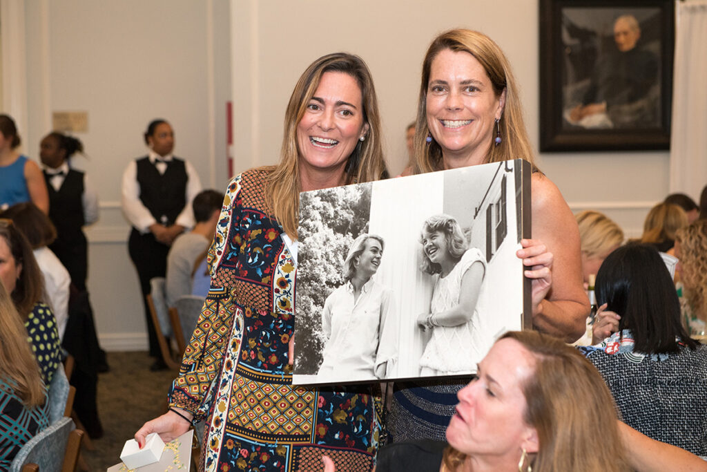 Two women smiling at an indoor event hold a black-and-white photo of a man and woman laughing. People are seated at tables in the background.