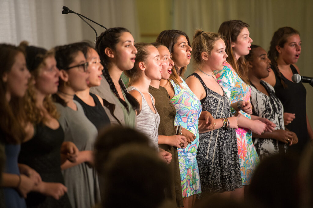 A group of young women stands in a line, singing together on stage. They hold hands or link arms and sing into a microphone, with expressions of focus and emotion.
