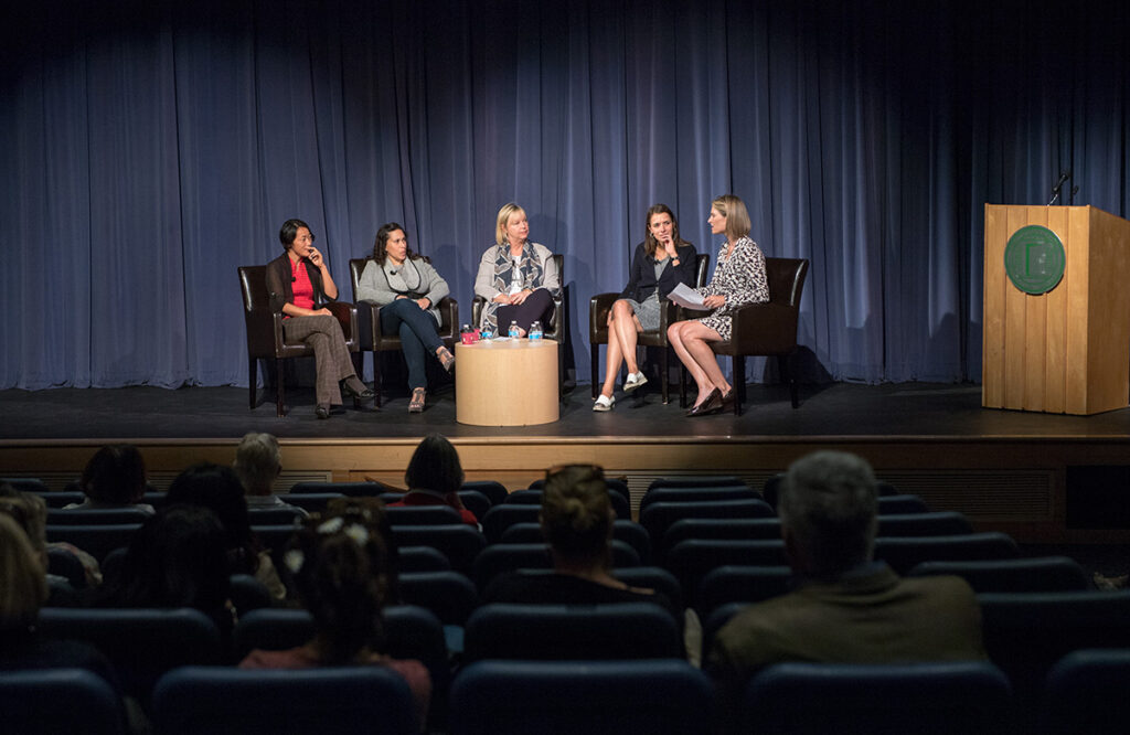 Five women sit on stage in a panel discussion. Four are seated on chairs, one on a stool, with a round table in the center. An audience watches from theater seats. Blue curtains hang in the background.