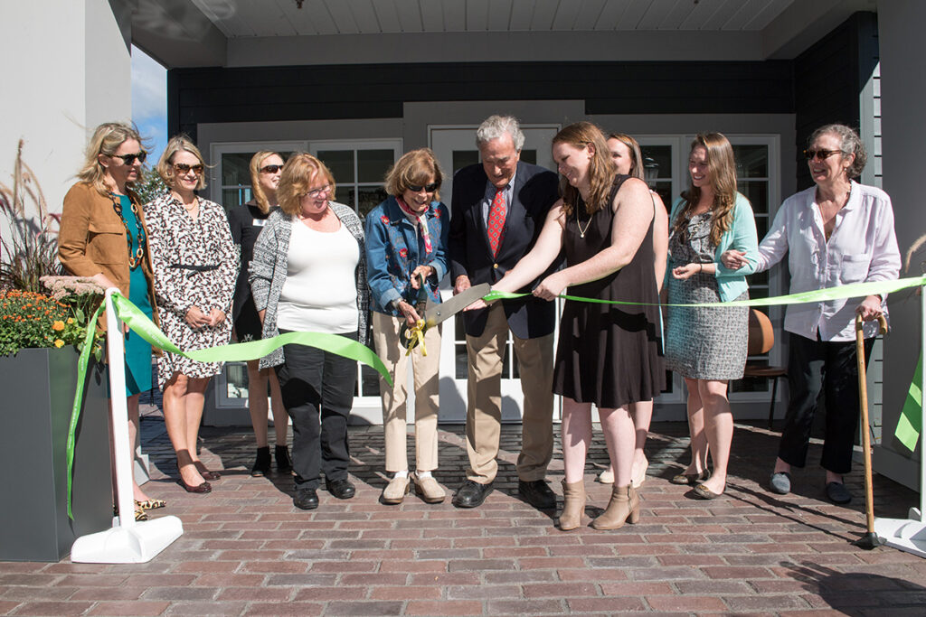 A group of people stand outside a building, smiling as a woman cuts a green ribbon with large scissors during a ribbon-cutting ceremony on a sunny day.