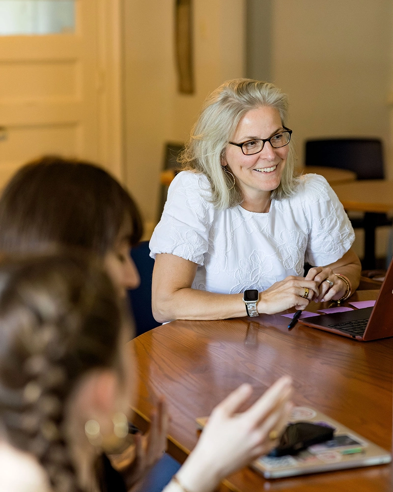 A woman with glasses and a white shirt smiles while sitting at a table with a laptop, talking to two people whose faces are out of focus in the foreground.