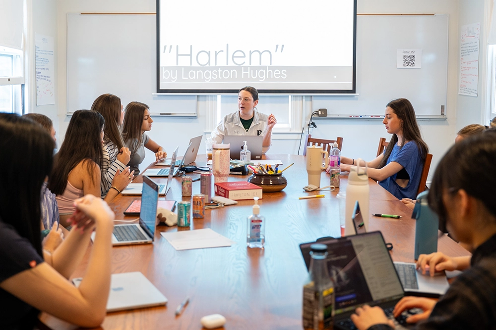 A group of students sit around a table with laptops, listening to a teacher. A screen displays Harlem by Langston Hughes. The setting appears to be a classroom or study group.