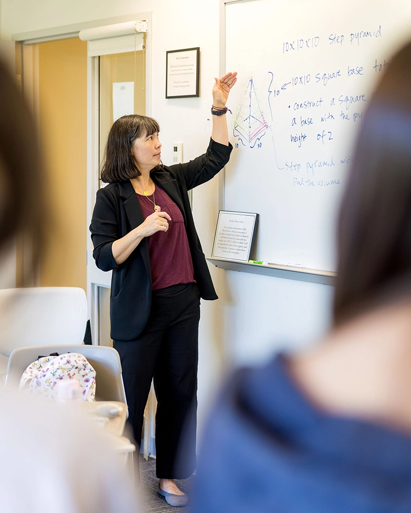 A woman stands by a whiteboard, pointing to a geometric diagram and handwritten math notes, engaging with students in a classroom setting.