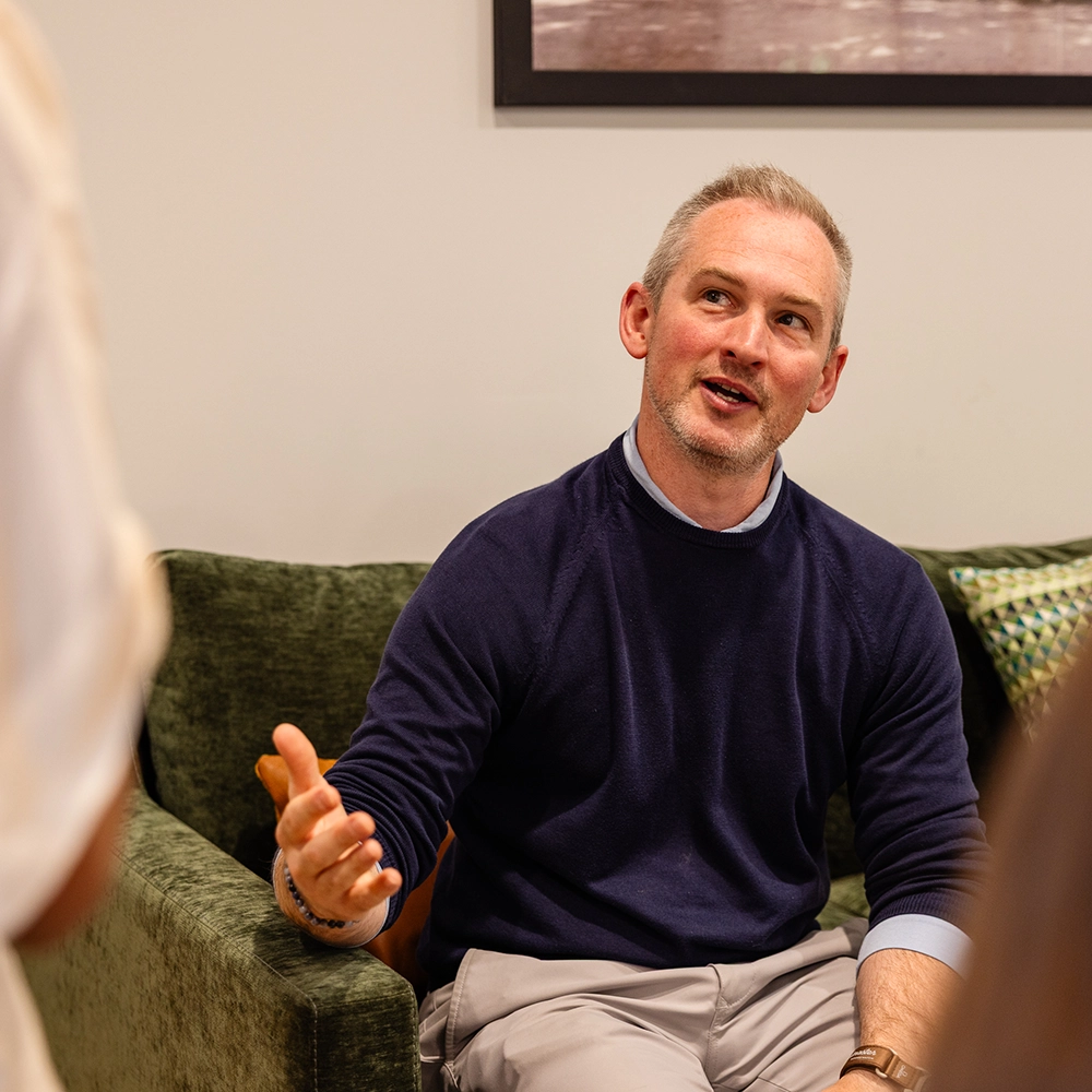 A man with short gray hair and a navy sweater sits on a green couch, gesturing with his hand and speaking to someone out of frame. A piece of patterned decor and part of a framed picture are visible behind him.
