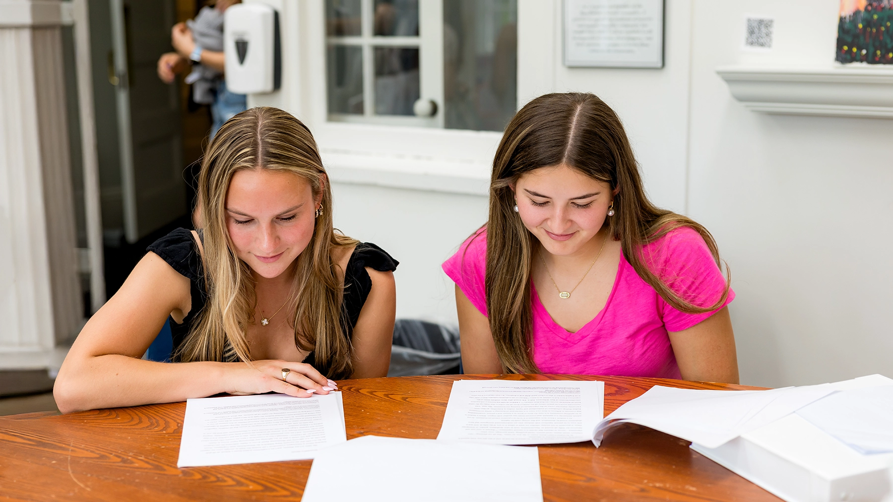 two high school girls sit at a table together reading some papers