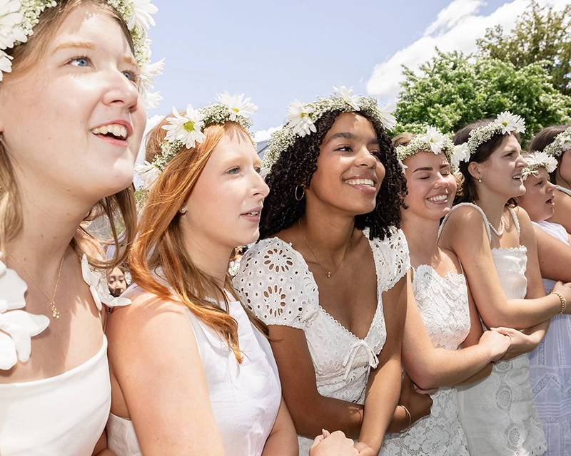 A group of young women in white dresses and flower crowns stand outdoors, smiling and linking arms, with greenery and a partly cloudy sky in the background.