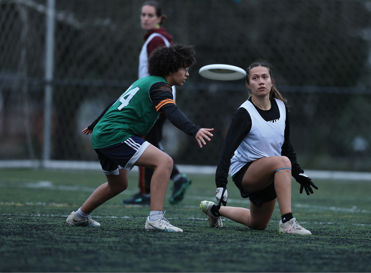 Two young players compete in an ultimate frisbee game; one kneels to catch the frisbee while the other defends. A goalie stands in the background near the net.
