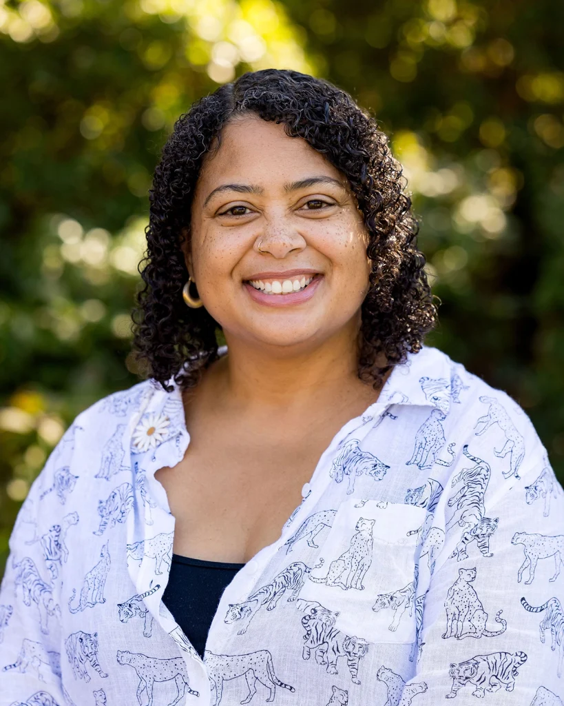 A woman with curly hair, wearing hoop earrings and a white shirt with animal prints, smiles warmly outdoors with greenery in the background.