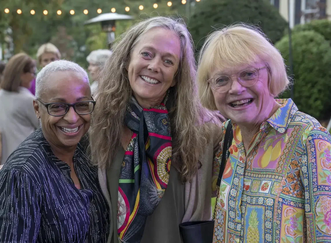 Three smiling Ancients pose together outdoors at an event, dressed in colorful clothing. String lights and greenery are visible in the background.