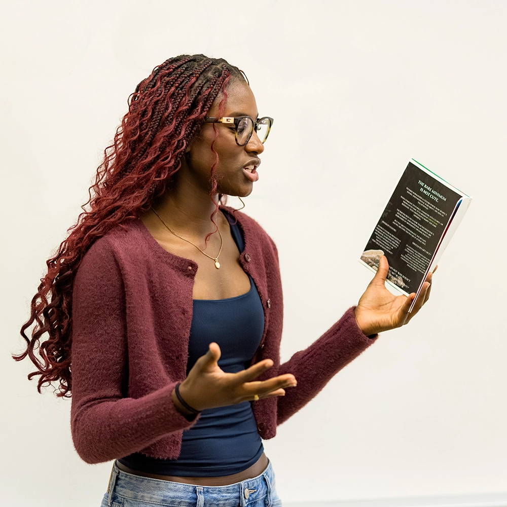 A woman with long braided hair and glasses, wearing a maroon sweater and black top, speaks while holding up a book in her right hand against a plain white background.