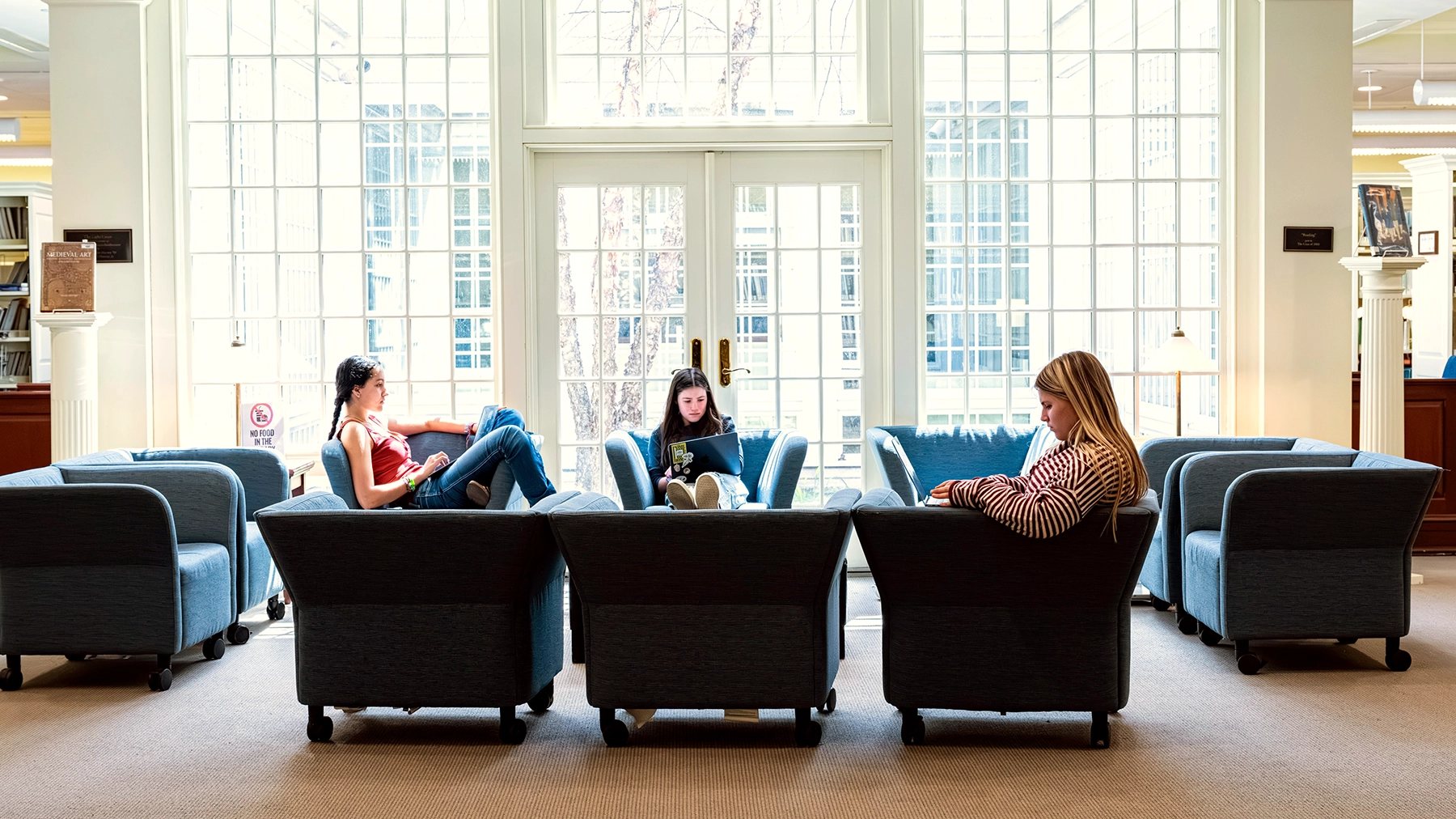 Three people sit on blue armchairs in a bright room with large windows, reading or using laptops. Bookshelves and more chairs are visible in the background.