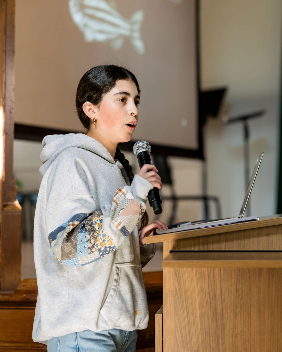 A young person in a gray hoodie speaks into a microphone at a podium, with a laptop in front of them and a projection screen with a fish image in the background.
