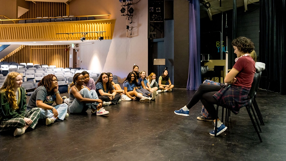 A group of students sit on a stage floor in a theater, facing two seated people who appear to be leading a discussion or workshop. The background shows rows of empty seats.